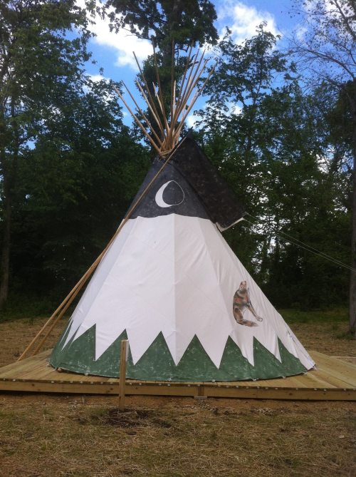 A white and green teepee with a black top, featuring a moon design and a painted animal, set in a wooded area.