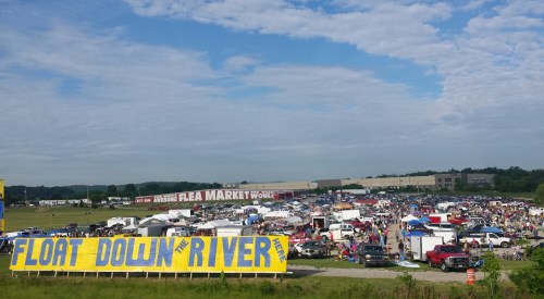 A bustling flea market scene with numerous tents and vehicles, set against a blue sky and a large sign reading "FLOAT DOWN THE RIVER."