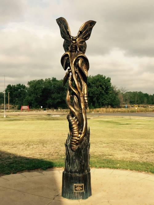 A tall bronze sculpture of a butterfly entwined with a serpent, set against a cloudy sky and grassy landscape.