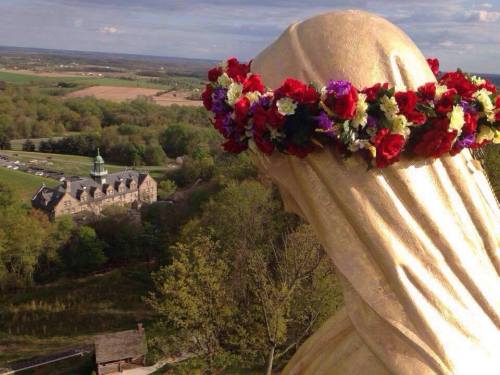 A golden statue of a figure wearing a floral crown overlooks a lush landscape and a building below.