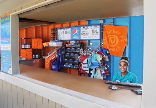 A beach kiosk with a smiling employee, colorful towels, and a menu board displaying drinks and snacks.