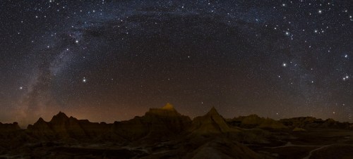 A panoramic view of a starry night sky over rugged, illuminated rock formations in a desert landscape.