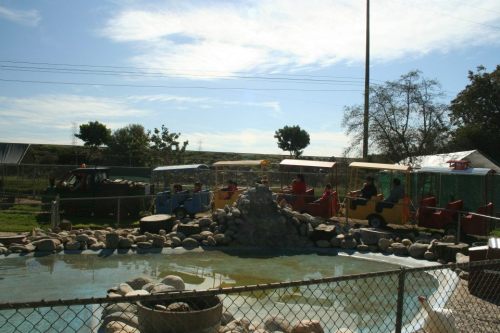 A colorful amusement ride area with a small pond, surrounded by a fence and trees under a clear blue sky.