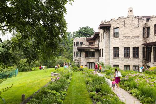 A historic stone mansion surrounded by lush gardens, with people walking along a path on a cloudy day.
