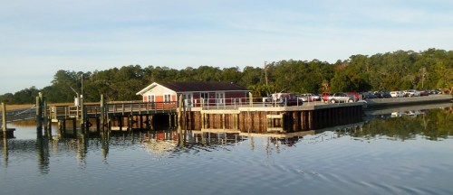 A wooden pier with a red building, surrounded by calm water and trees, reflecting the scene in the water.