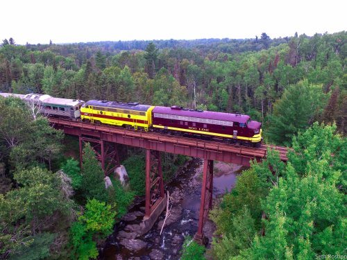 A vintage train crosses a wooden trestle bridge over a river, surrounded by lush green trees.