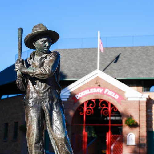 A bronze statue of a baseball player holding a bat stands in front of Doubleday Field's entrance.