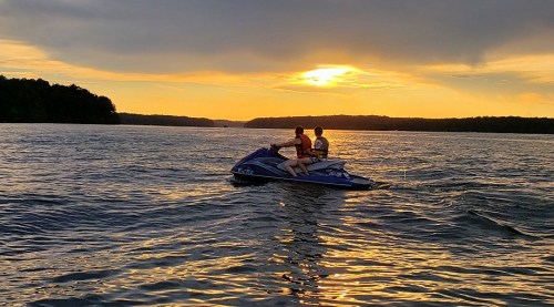 Two people ride a jet ski on a lake at sunset, with golden light reflecting on the water.