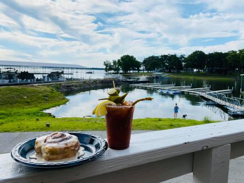 A cinnamon roll and a cocktail sit on a table overlooking a serene lake with boats and a person in the distance.