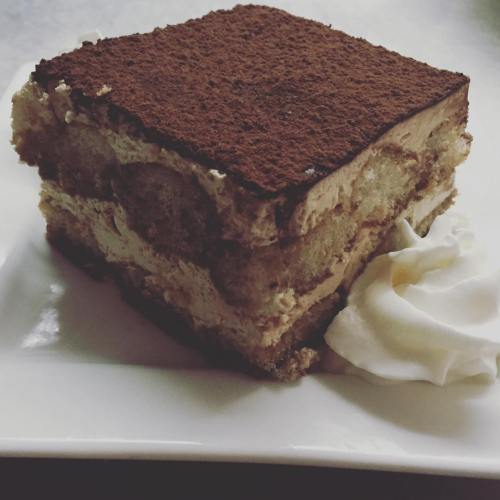 A slice of tiramisu topped with cocoa powder, served on a white plate with a dollop of whipped cream.