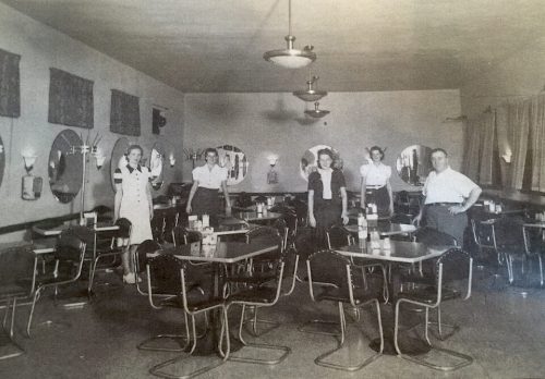 A vintage diner interior with five people standing among empty tables and mirrors on the walls.