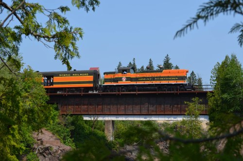 A vintage train crosses a bridge surrounded by trees, showcasing a scenic landscape under a clear blue sky.