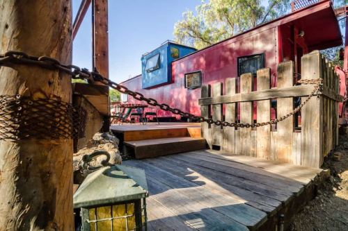 A rustic wooden deck with a chain fence, leading to a red train car surrounded by trees and sunlight.
