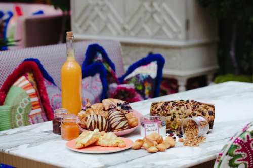 A table set with various pastries, a bottle of juice, and jars of spreads, surrounded by colorful cushions.