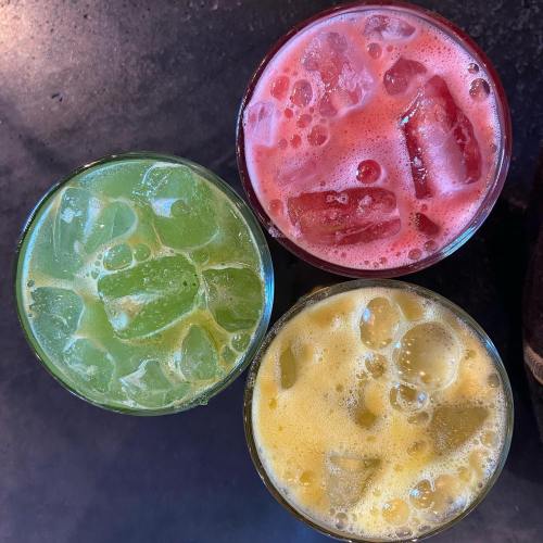 Three colorful drinks in glasses: green, red, and yellow, all filled with ice and bubbles on a dark surface.