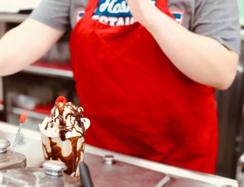 A person in a red apron prepares a sundae topped with whipped cream, chocolate sauce, and cherries.