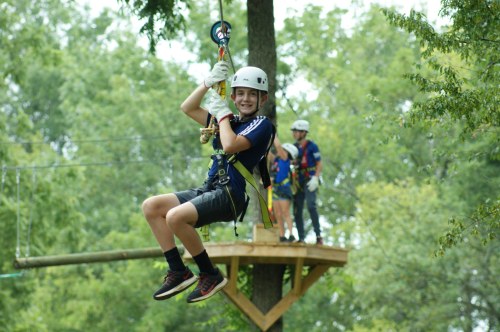 A young boy in a helmet zip lines through a forest, while others prepare on a platform in the background.