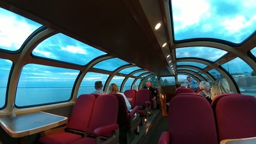 Interior of a train car with large windows, featuring red seats and passengers enjoying a scenic view of the water.