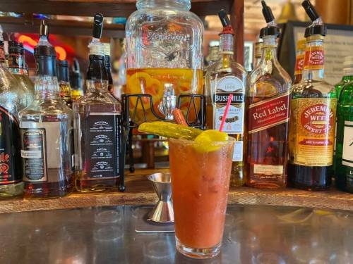 A glass of Bloody Mary garnished with a pickle, set against a backdrop of various liquor bottles on a bar.