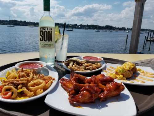 A sunny outdoor table with plates of fried calamari, chicken wings, and a taco, alongside a bottle of cucumber vodka.