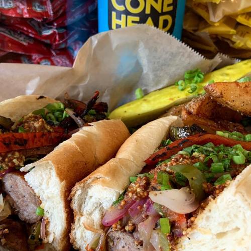 Three loaded sausage sandwiches topped with grilled vegetables and green onions, with a pickle and a drink in the background.