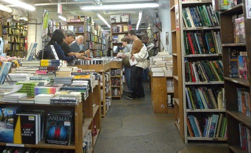 A busy bookstore aisle with customers browsing books on shelves and tables, surrounded by stacks of various titles.
