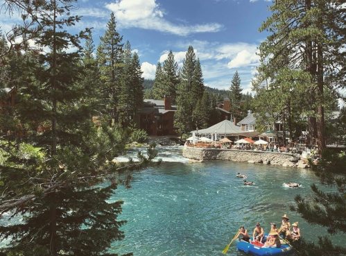 A scenic river surrounded by trees, with people kayaking and relaxing by a riverside restaurant on a sunny day.
