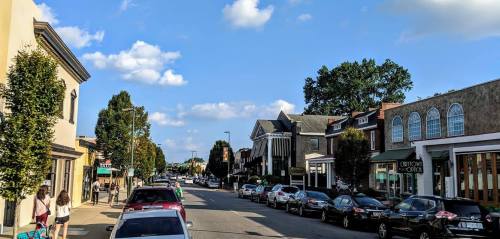 A sunny street scene with shops and parked cars, lined with trees under a blue sky with scattered clouds.