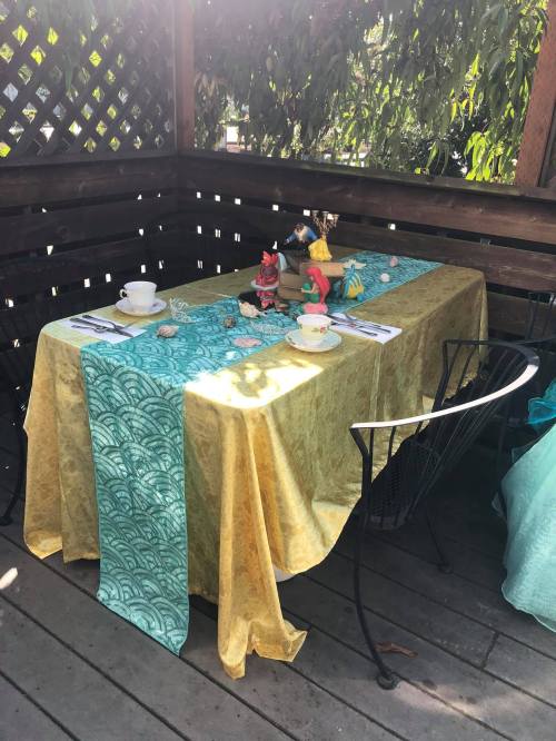 A table set outdoors with colorful tablecloths, decorative figurines, and tea cups, surrounded by greenery.