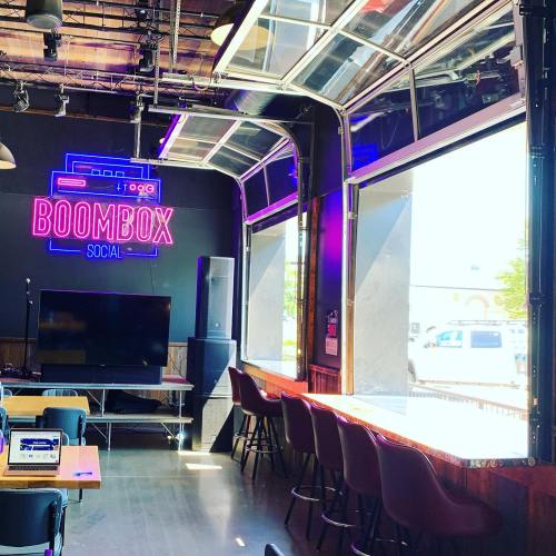 Interior of a modern venue with a neon "BOOMBOX" sign, large TV, and bar seating by open windows.
