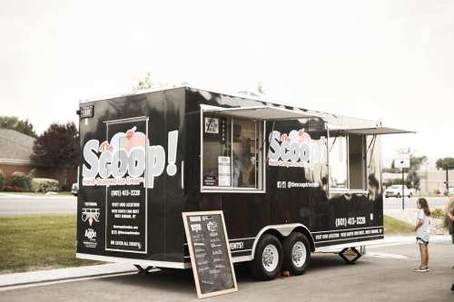 A black ice cream truck named "The Scoop!" with a menu board outside and a child standing nearby.