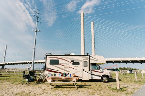 An RV parked by a bridge, with a table set up outside and a scenic view of the sky and grass.