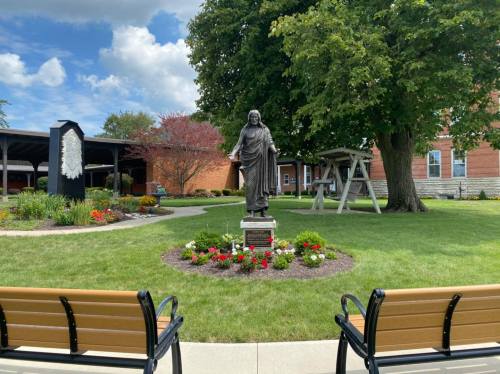 A statue of Jesus stands in a garden surrounded by flowers, with benches and a building in the background under a blue sky.