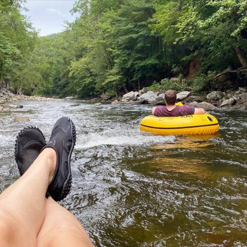 A person relaxes in a river on a yellow inner tube, with their feet visible in the foreground and trees lining the banks.