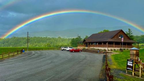 A rustic building with a rainbow arching over it, set against a backdrop of green hills and a cloudy sky.