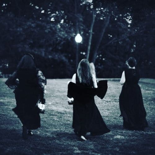 Three women in black dresses walking away in a dimly lit park, surrounded by trees.