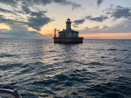 A lighthouse stands on a small island in the water at sunset, with colorful clouds reflecting in the waves.