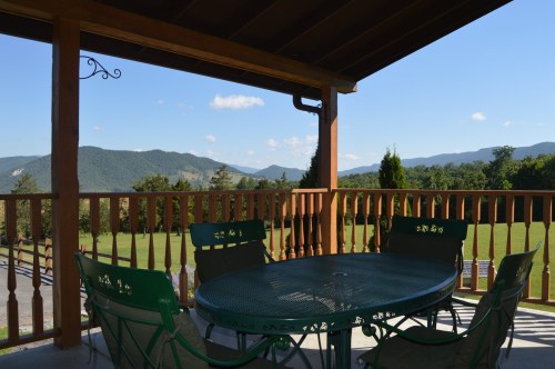 A scenic view from a porch with a round table and chairs, overlooking green mountains and a clear blue sky.