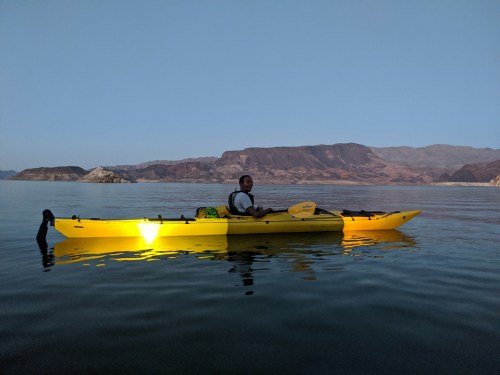 A person sits in a bright yellow kayak on calm water, surrounded by mountains under a clear sky.