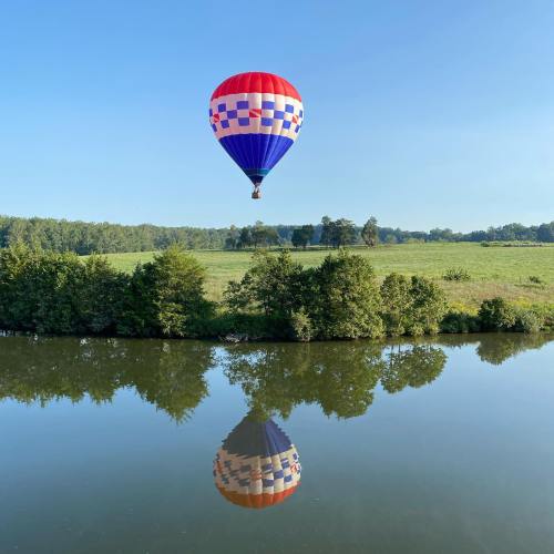 A colorful hot air balloon floats above a calm lake, reflecting on the water, surrounded by green trees and blue sky.