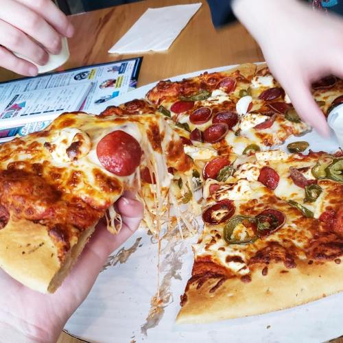 A hand pulls a slice of pepperoni and jalapeño pizza, with melted cheese stretching from the pie on a wooden table.