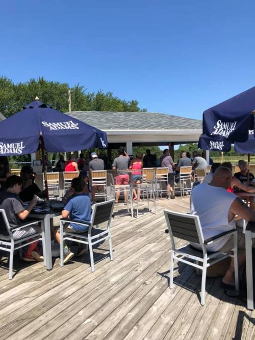 Outdoor bar scene with patrons seated at tables under umbrellas, enjoying a sunny day. Green trees in the background.