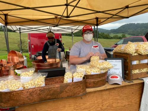 A popcorn stand at an outdoor market, with two vendors wearing masks and various popcorn flavors displayed.