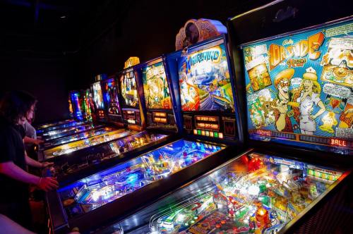 A row of colorful pinball machines with players engaged, featuring vibrant lights and retro designs.
