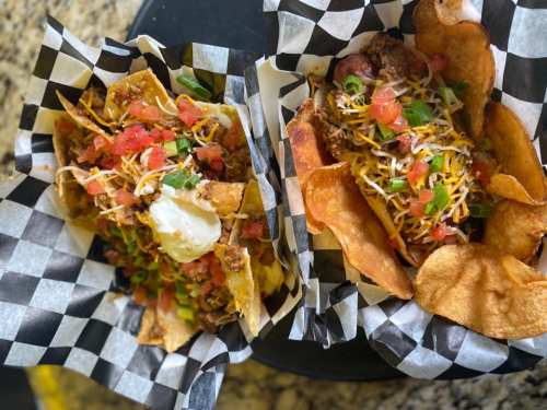 Two baskets of nachos topped with cheese, sour cream, tomatoes, and green onions, served on a checkered paper.