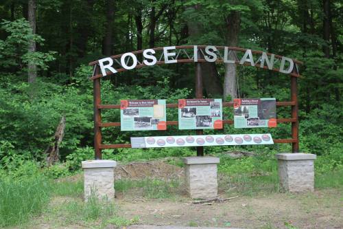 Sign for Rose Island with informational panels, surrounded by trees and greenery.