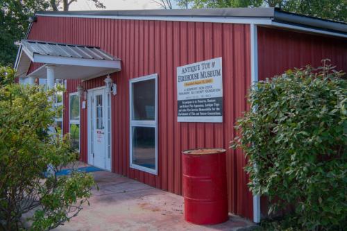 Exterior of the Antique Tot & Firehouse Museum, featuring a red building with a sign and greenery in front.