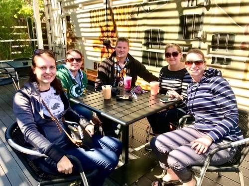 A group of five people sitting at a table outdoors, enjoying drinks and chatting in a sunny setting.