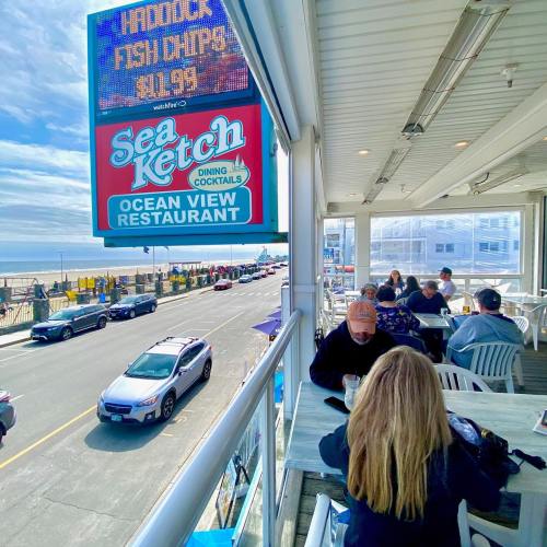 A seaside restaurant with outdoor seating, diners enjoying meals, and a view of the beach and ocean.