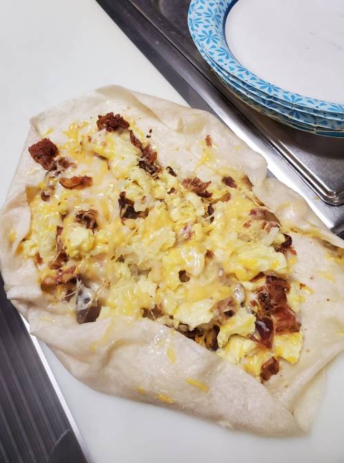 A large burrito filled with scrambled eggs, cheese, and bacon, resting on a countertop next to a paper plate.